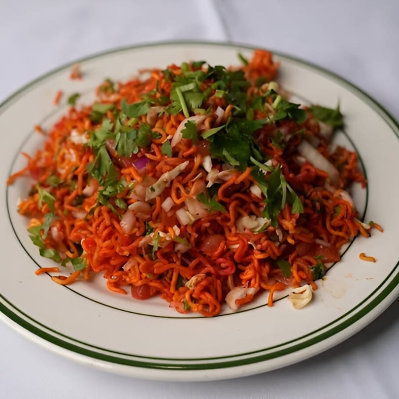 A white plate with green trim holds a serving of orange Indian noodles, garnished with cilantro and onions.