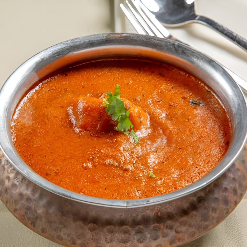 A copper bowl filled with vibrant orange Indian curry, garnished with fresh cilantro.