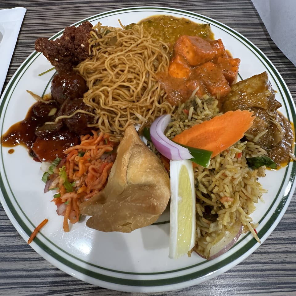 A white plate filled with various Indian dishes, including noodles, paneer, rice, samosa, and curries.