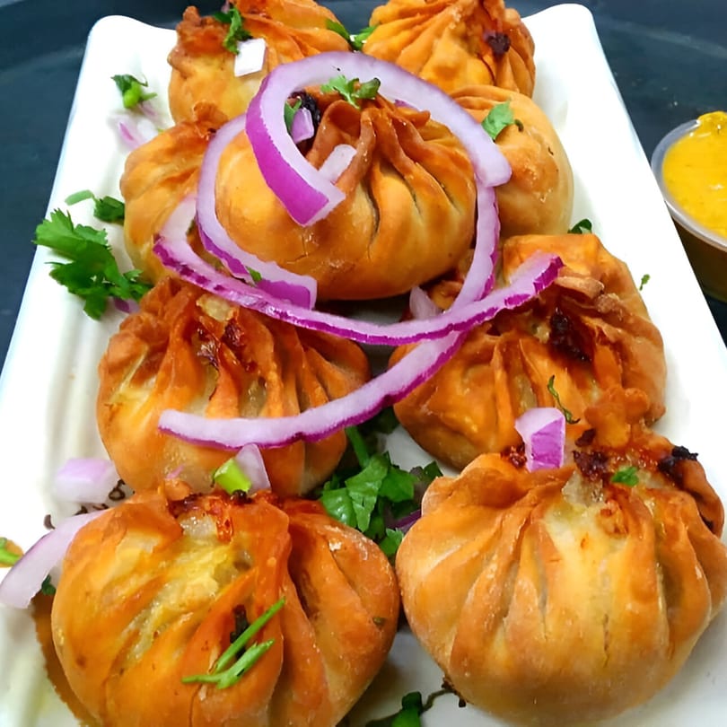 Fried momos, garnished with red onion and cilantro, on a white plate with a side of yellow sauce.