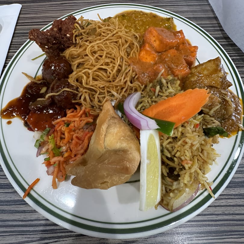 A white plate with green stripes filled with various Indian dishes including noodles, rice, samosa, and curries.